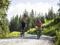 Ein Hotel in Großarl für Aktive | für Genießer | für dich Zwei Radfahrer fahren auf einem Waldweg mit Seilbahn und Tannen im Hintergrund